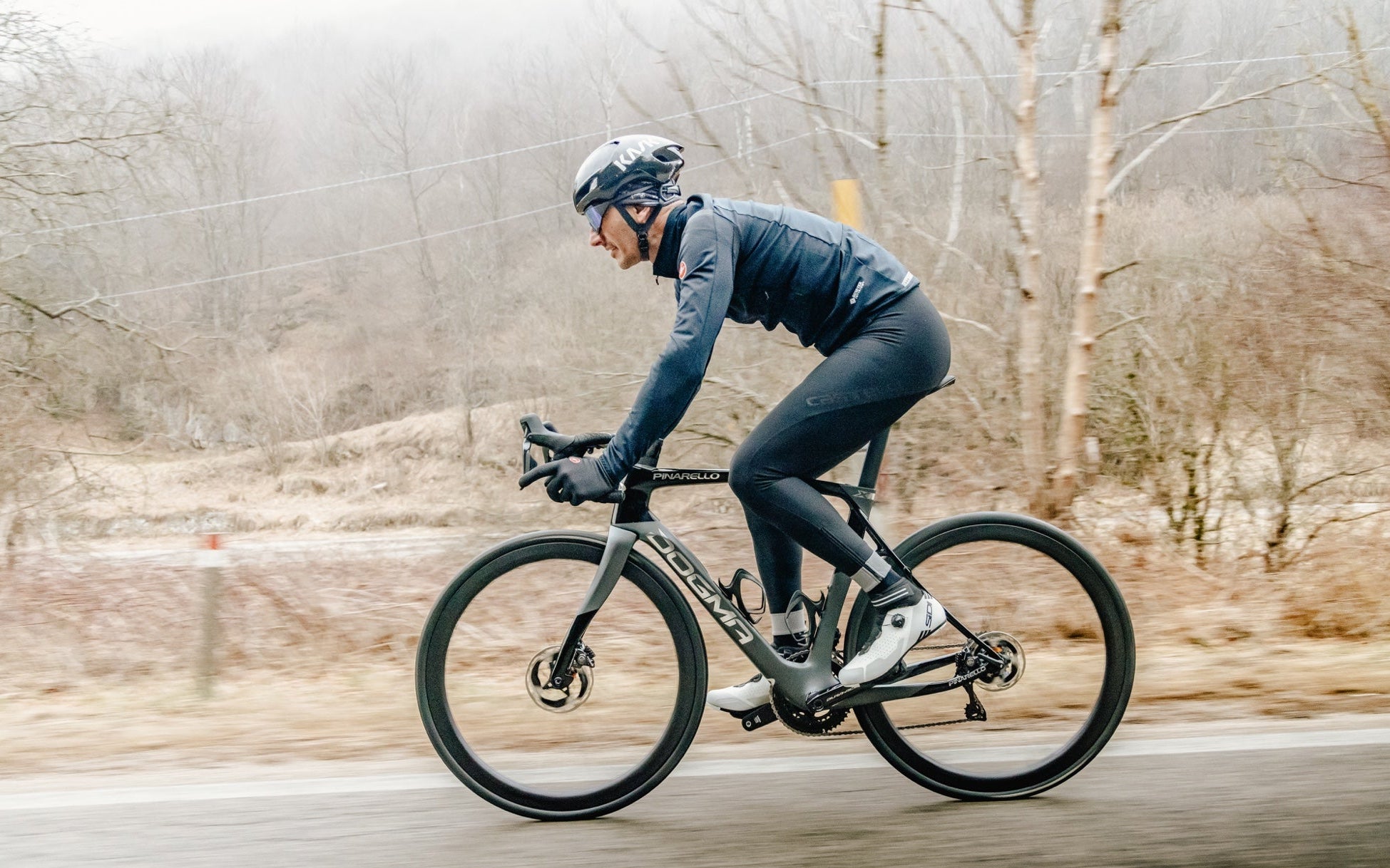 Cyclist riding through snowy conditions on a road with bare trees in the background
